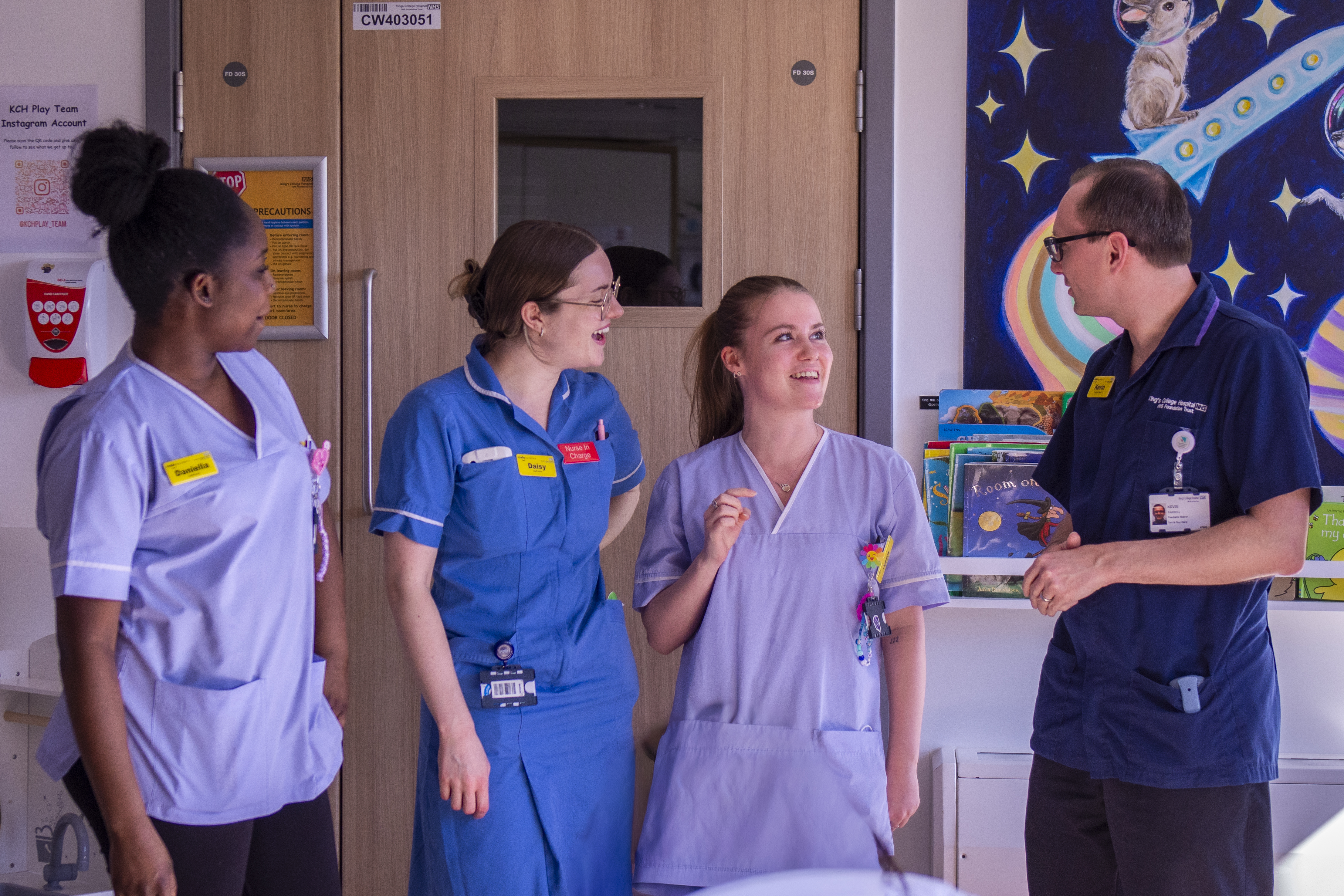 Four King’s healthcare staff members stand together in a newly refurbished Playroom, funded by the Charity, at King’s College Hospital. Three wear light blue or purple medical uniforms and one wears dark blue scrubs. A colourful children's space-themed mural and books are visible in the background, creating a friendly, paediatric setting.